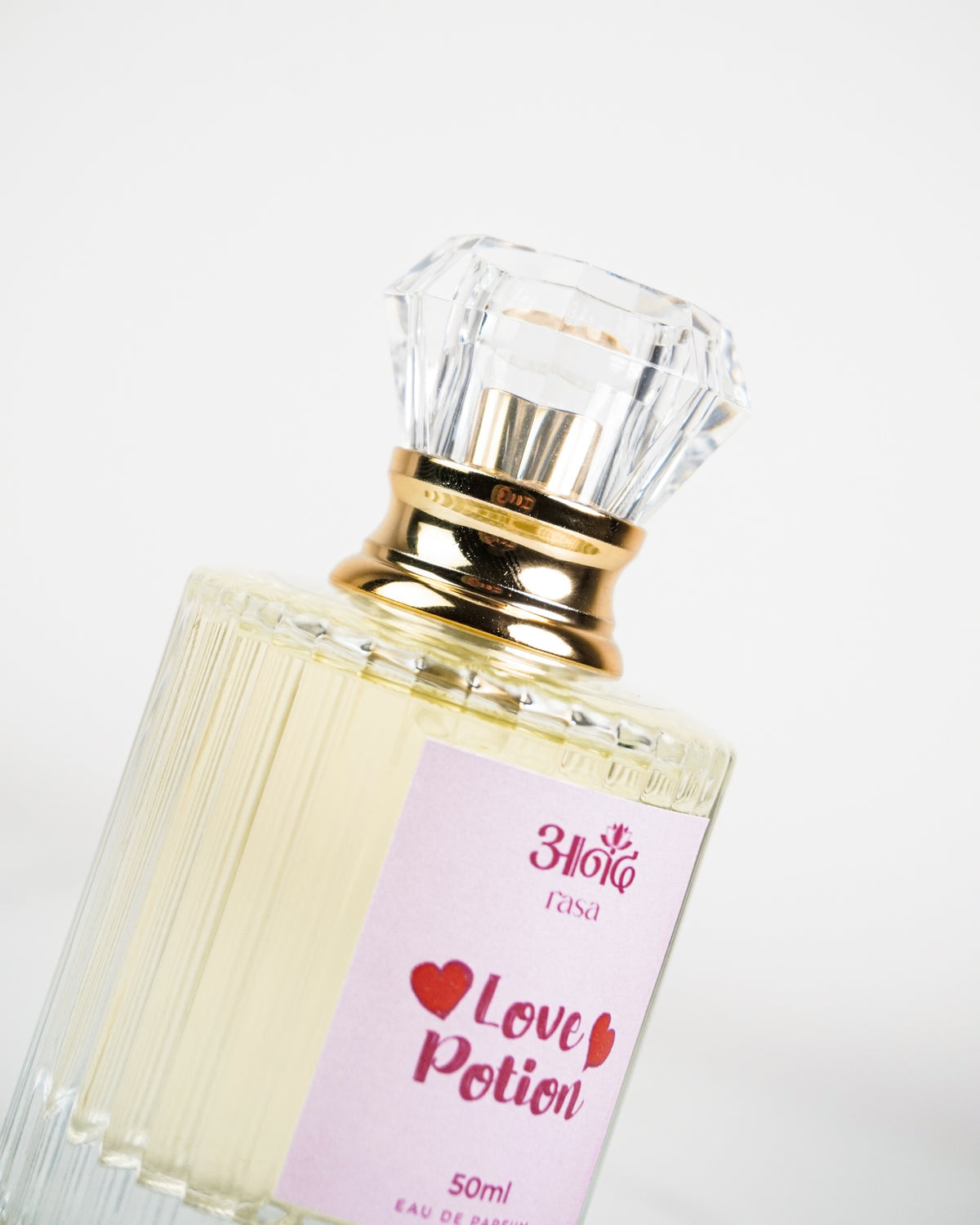 Love Potion 50ml perfume in a fluted clear glass bottle with a pink label, gold collar and faceted transparent cap on a white marble-style background.